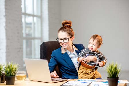 Young multitasking businessmam dressed in the suit working with laptop and documents sitting with her baby son at the officeの写真素材