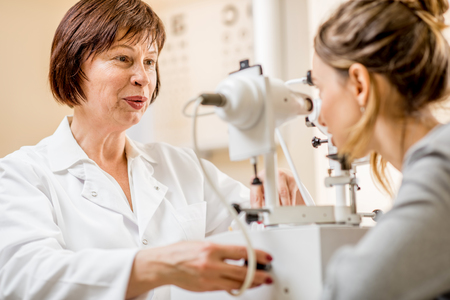 Senior woman ophthalmologist checking vision with laser device to the young female patient sitting in the officeの写真素材