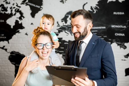 Happy mother and baby boy choosing tour for a summer vacation with male agent at the travel agency office with world map on the background. Image focused on a babyの写真素材