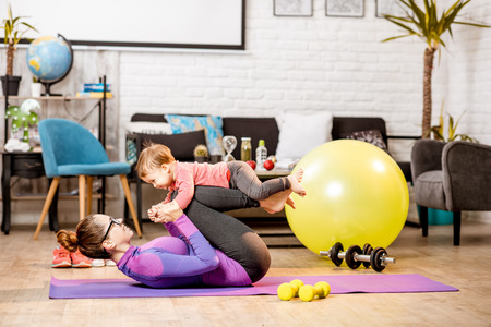 Young mother in sportswear doing exercise lifting with legs her baby son on the mat at homeの写真素材