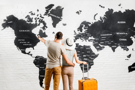 Young couple of travelers standing back near the big world map on the background choosing a place...