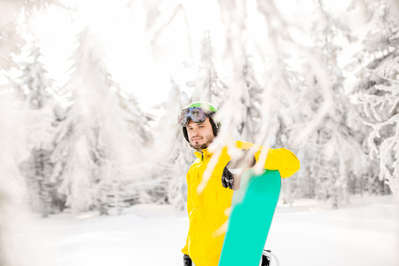 Portrait of a man in winter clothes standing with snowboard in the beautiful snow-covered fir forestの写真素材