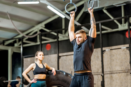 Handsome man in black sportswear pulling up on the gymnastic rings with woman on the background in...