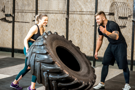 Athletic woman pushing a big tire training with a handsome trainer in the gymの写真素材