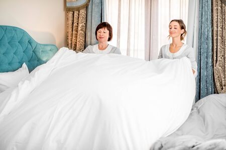 Senior chambermaid with young assistant in uniform making a bed in the luxury hotel bedroomの写真素材