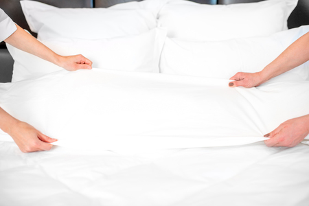 Two chambermaids in uniform making a bed with clean bedclothes in the luxury hotel bedroom. Close-up view on the hands and bedclothesの写真素材