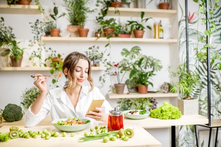 Young woman eating healthy food sitting with smartphone in the beautiful interior with green flowers on the backgroundの写真素材