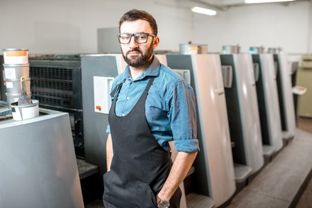 Portrait of a handsome typographer standing at the printing manufacturing with offset machine on the backgroundの写真素材