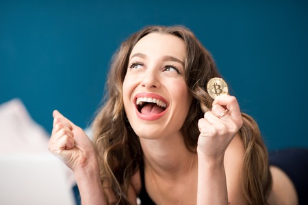 Happy young woman with excited emotions holding crypto coin lying with laptop at homeの写真素材