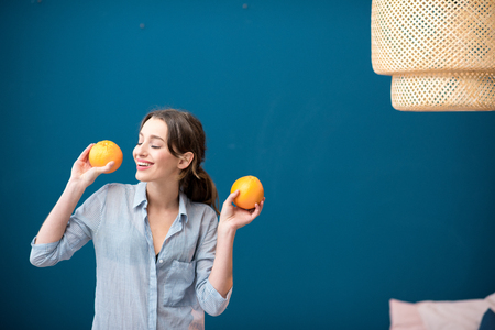 Portrait of a young and happy woman with oranges on the blue wall background at homeの写真素材
