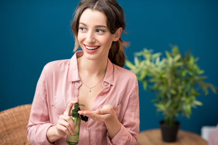 Portrait of a young woman holding green bottles with lotion on the blue wall background at homeの写真素材