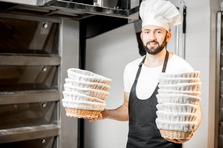 Portrait of a baker in uniform standing with baking forms near the oven in the bakery manufacturing