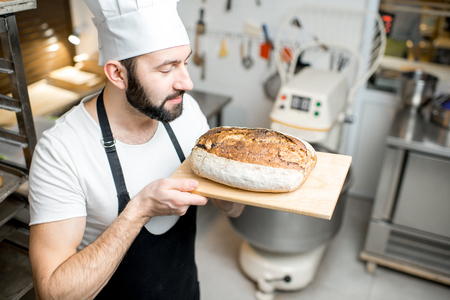 Portrait of a handsome baker in uniform standing with fresh bread in the small bakery manufacturingの写真素材