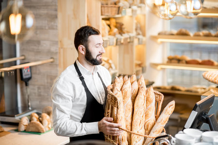 Handsome bread seller with basket full of baguettes in the beautiful store with bakery productsの写真素材