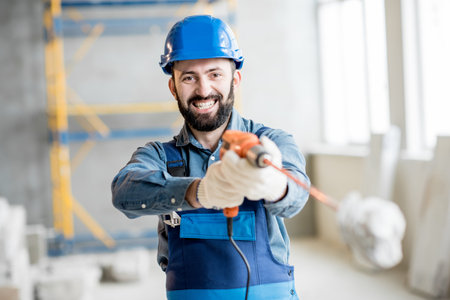 Portrait of a builder in working uniform standing with drill at the construction site indoors