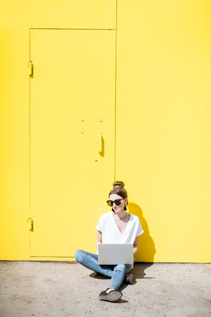 Young woman working with laptop sitting on the yellow wall background outdoorsの写真素材