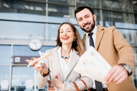 Portrait of a happy business couple in coats stading with tickets and toy airplane in front of the airport entranceの写真素材