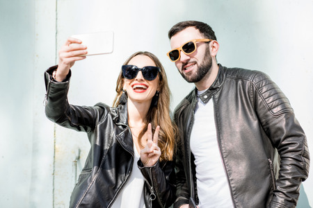 Young stylish couple in leather jackets making selfie with phone on the light green wall backgroundの写真素材
