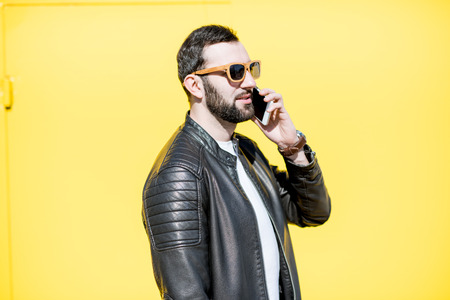 Portrait of stylish man in leather jacket talking with phone on the yellow background outdoorsの写真素材