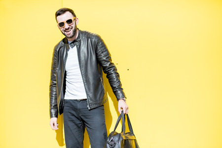 Portrait of stylish man in leather jacket with bag on the yellow background outdoorsの写真素材