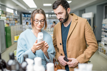 Young couple dressed in coats choosing medicine standing with box in the pharmacy storeの写真素材