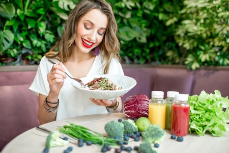 Young and beautiful woman enjoying salad sitting at the table with smoothies and healthy food ingredients on the green wall backgroundの写真素材
