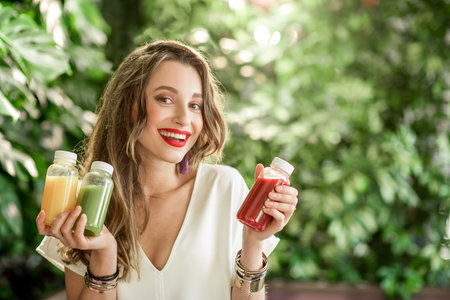 Portrait of a young happy woman holding bottles with healthy smoothie drinks standing on the green...