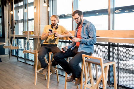 Two men dressed casually using smartphones sitting near the window of the modern cafe interiorの写真素材