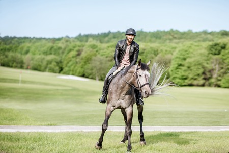 Handsome man in leather jacket with protective helmet riding a horse on the green meadowの写真素材