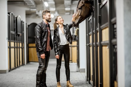 Young couple riders in leather jackets standing in the beautiful stable stroking a horse headの写真素材
