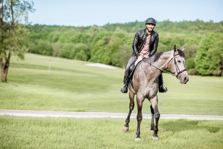 Handsome man in leather jacket with protective helmet riding a horse on the green meadowの写真素材