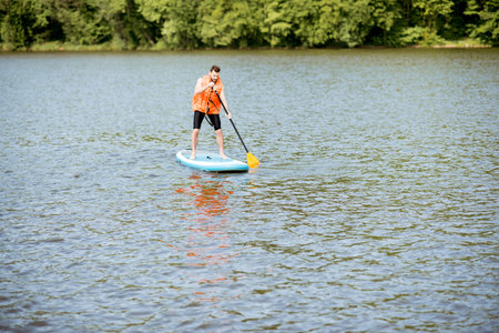 Man in life vest swimming on the standup paddleboard on the lake near the green forest