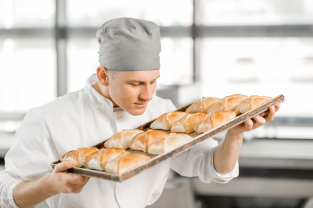 Portrait of a handsome baker with tray full of freshly baked buns standing at the bakery manufacturingの写真素材