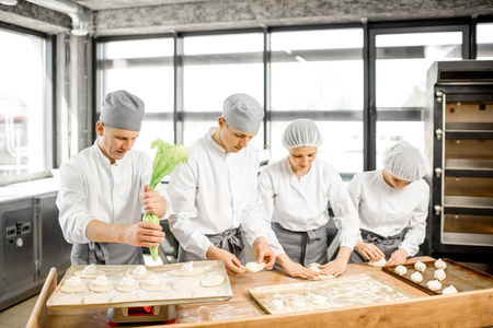 Group of young workers in uniform filling buns for baking on the wooden table standing together at the modern manufacturingの写真素材