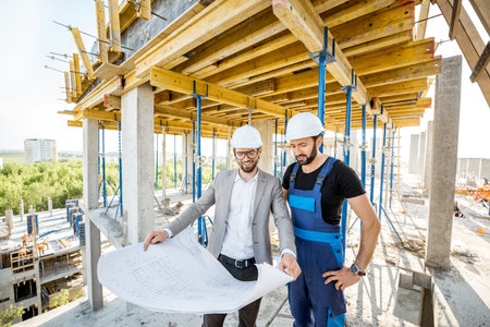 Engineer standing with builder supervising the construction process with house drawings on the...