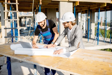 Engineer with worker in uniform working with architectural drawings and laptop at the table on the construction site outdoorsの写真素材