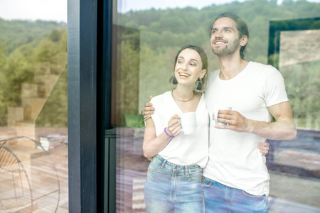 Young and lovely couple standing together looking outside the window at home, view through the...