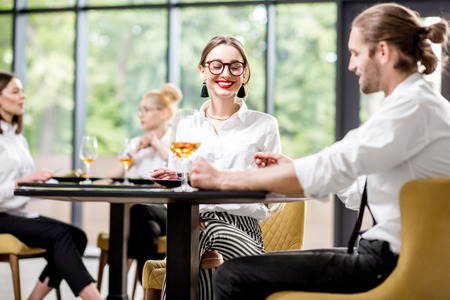 Beautiful couple clinking with wine glasses during a business lunch with delicious meals at the modern restaurant with business people on the backgroundの写真素材