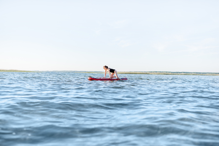Beautiful young woman in black swimsuit paddleboarding on the lake with waves during the morning lightの写真素材