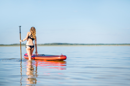 Beautiful woman in black swimsuit standing with paddleboard in the calm water with reflection during the sunset lightの写真素材
