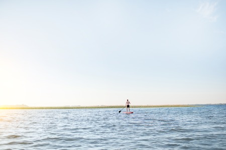 Man paddleboarding on the lake during the morning light, wide landscape view with blue water and skyの写真素材