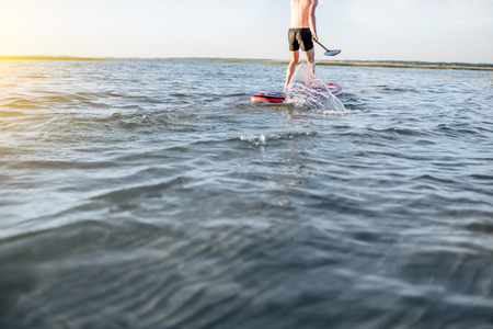 Man rowing with paddleboard on the lake with blue water during the sunrise, cropped image without mans faceの写真素材