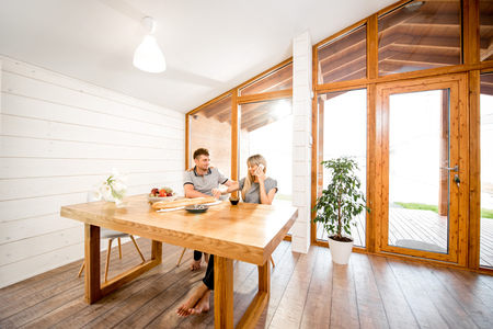 Young and happy couple having a breakfast sitting at the wooden table in the modern country house with big window on the backgroundの写真素材