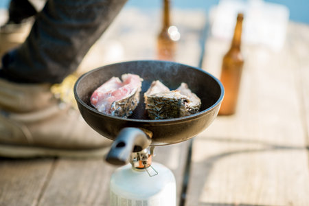 Frying two fish steakes on the burner during the picnic with beer and fishing tackles outdoors