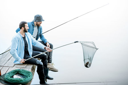 Two friends catching fish with fishing net and rod sitting on the wooden pier at the lakeの写真素材