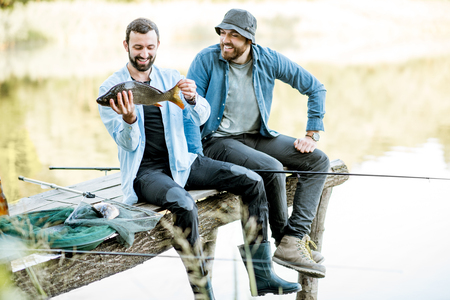 Two happy fishermen holding caught fish sitting on the wooden pier during the fishing on the lake at the morningの写真素材