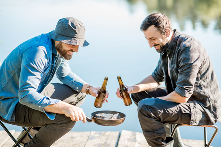 Two fishermen frying fish sitting with beer during the picnic on the wooden pier near the lake in the morningの写真素材