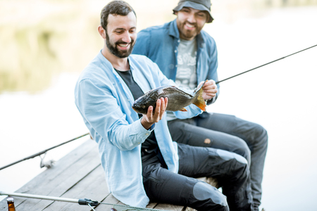 Two happy fishermen holding caught fish sitting on the wooden pier during the fishing on the lake at the morningの写真素材