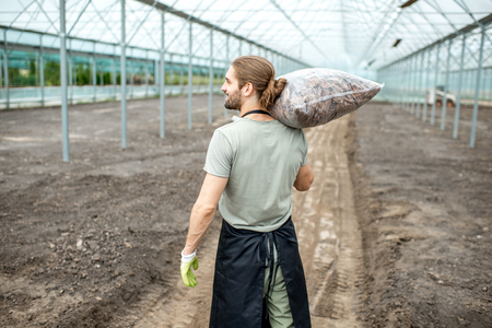 Handsome farmer in working uniform carrying bag with bark for mulching in the glasshouseの写真素材
