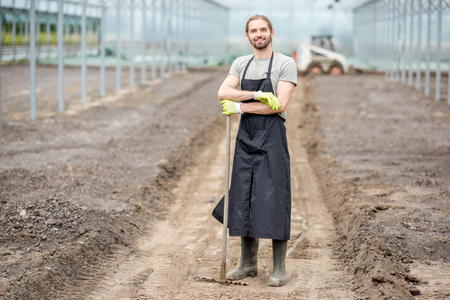 Portrait of a handsome farmer in working uniform standing with rake in the glasshouseの写真素材